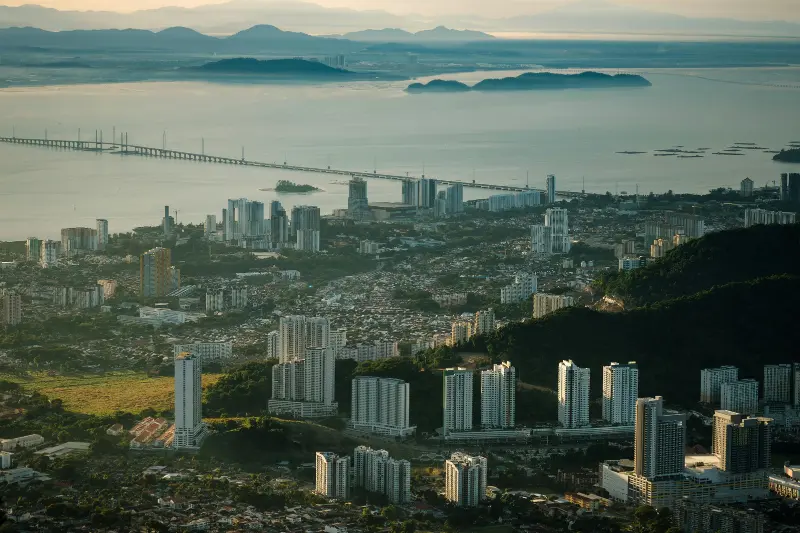 High-angle aerial view of the Penang Bridge spanning across the sea towards mainland Malaysia, showcasing the Gelugor waterfront skyline and lush green hills.